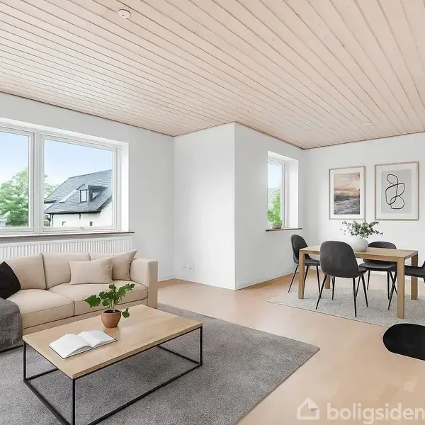 En stue har en rund spisebord med stole og en vase, placeret foran store vinduer, der giver udsigt til grønne omgivelser. En sofa med planter bagved står i hjørnet.