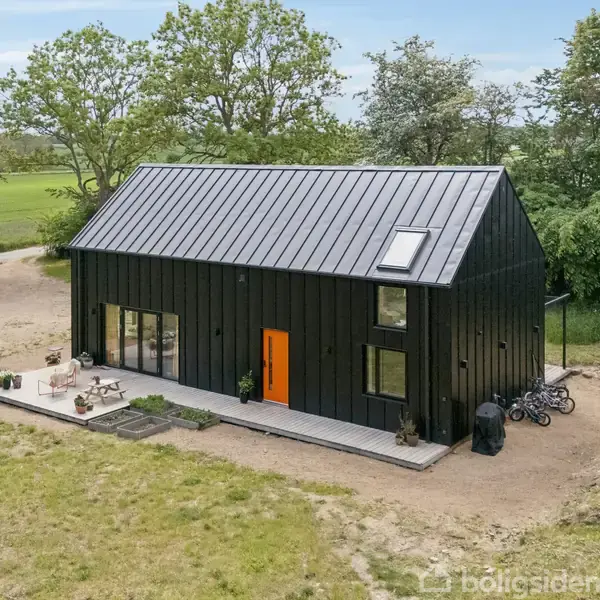 Sort hus med orange dør står på grusbelagt område. Huset har et skråt tag med ovenlysvindue. Terrassen har havemøbler. Omgivet af grønne marker og træer. Cykler ses ved siden.