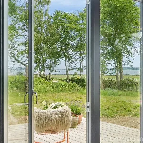 Glasdøre åbner mod en terrasse med en stol dækket af et tæppe. I baggrunden er der græsplæne og grønne træer med udsigt over vandet.