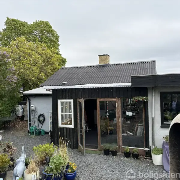 Et lille hus med sort træbeklædning står i en haveomgivet af træer og planter. Indgangen er enkel med flere blomsterkrukker. Himlen er overskyet.