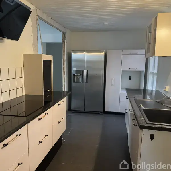 Et moderne køkken med sorte bordplader og hvide skabe. Et stort køleskab med dobbelt dør står i baggrunden ved siden af vasken. Der er flisebelagt backsplash.