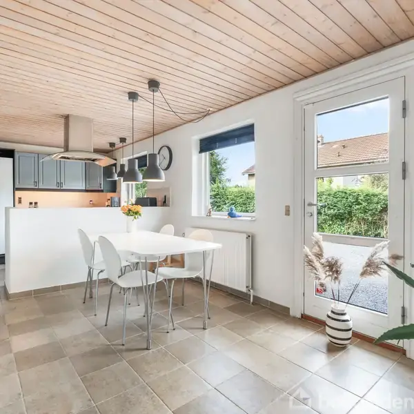 Et lyst køkkenalrum med et spisebord og fire stole midt i rummet. Køkkenet har grå skabe i baggrunden. En dør og et vindue byder på udsigt til en have.