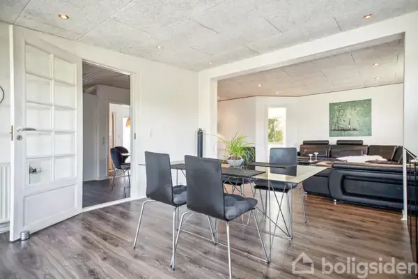 Et moderne spisestuebord med fire stole står på et mørkt trægulv. I baggrunden ses en stue med sorte sofaer og et kunstværk på væggen, med lyse vægge og lofter.