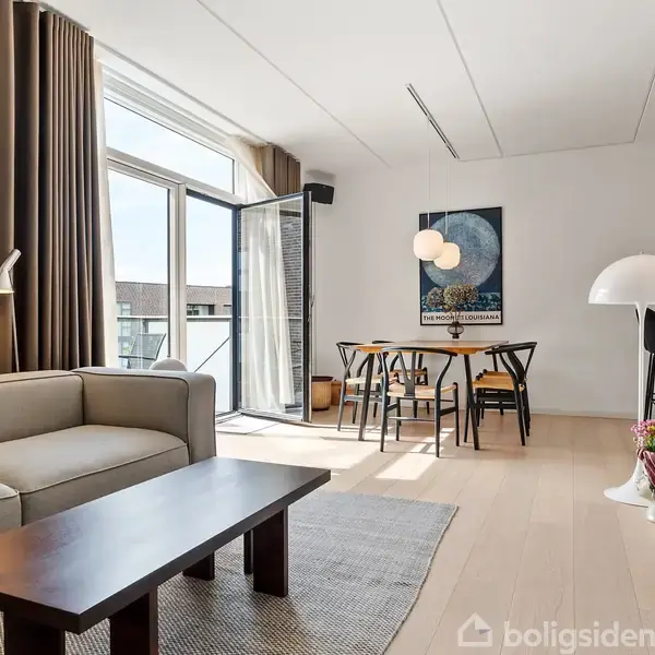 Et stilfuldt stueområde med en beige sofa ved et mørkt træbord. Sollys strømmer ind gennem store vinduer, og en spiseplads er synlig i baggrunden under en moderne lampe.