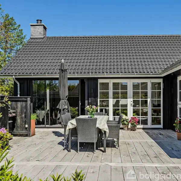 Sort træhus med hvide vinduer. Bord og stole står på en stor træterrasse med planter omkring. En åben parasol står ved siden af bordet. Blå himmel og grønne træer omkranser huset.
