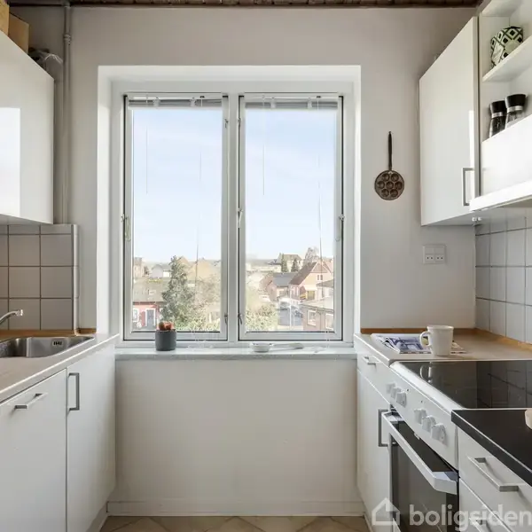 Køkken i hvidt design med moderne skabe og apparater. Udsigten gennem et vindue viser et bylandskab. En kop og kogebøger er på køkkenbordet.