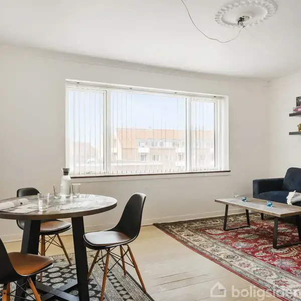 Rundt spisebord med sorte stole står i en lys stue. Blåt sofabord ved siden af mørk sofa. Tæpper dækker gulvet. Reoler med figurer hænger på væggen.