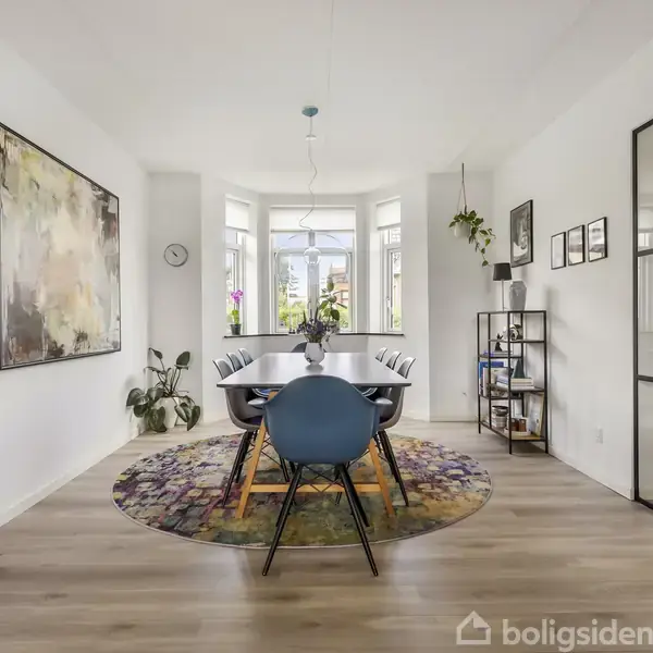 Spisebord med seks stole står centralt på rund tæppe, placeret i et lyst spisestueområde med stort abstrakt maleri, planter, reoler, og store vinduer, der lader naturligt lys komme ind.