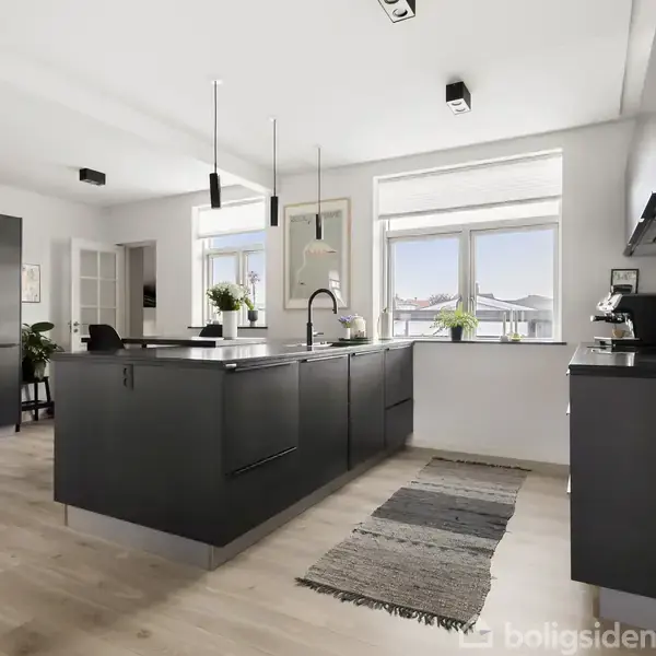 Et moderne køkken med mørke skabe og en central ø, hvorpå der står en vask. Rummet er lyst og rummeligt med store vinduer og lyse trægulve.