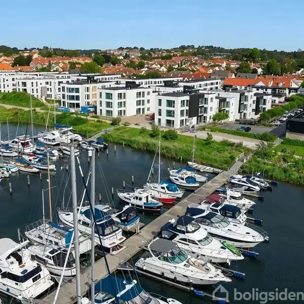 Både ligger fortøjet i en marina, omgivet af moderne bygninger og grønne områder. Flere både er fortøjet langs en bred kajplads, mens en by ses i baggrunden.