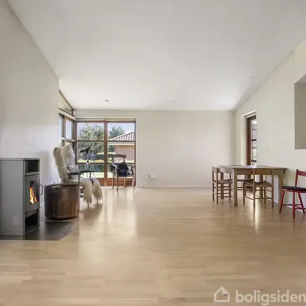 Et moderne, enkeltplans murstenshus står stille på en velplejet græsplæne, omgivet af en lav hæk og trådhegn. En blå himmel danner baggrund.