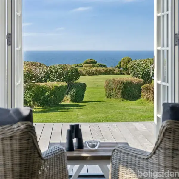 To kurvestole står på en terrasse foran åbne døre; de vender mod en velplejet græsplæne med havudsigt i det fjerne.
