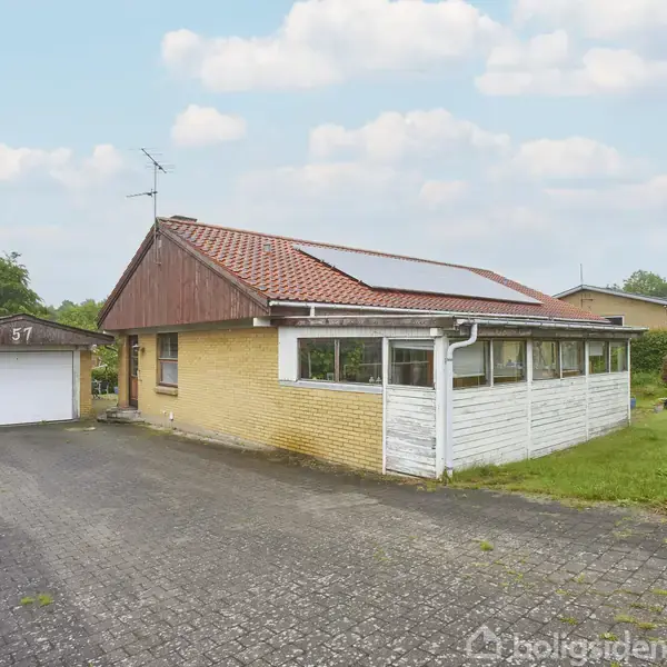 Et gult murstenshus med rødt tag har en tilstødende garage markeret "57". Solpaneler er installeret på taget. Huset ligger ved en brolagt indkørsel omgivet af grønne buske.