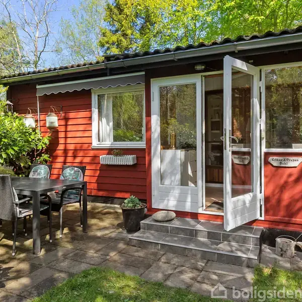 Rødt hus med åbne døre står i en solrig have. Foran står et havebord med stole omgivet af grønne træer og planter. Skilt ved døren siger "Colonien".