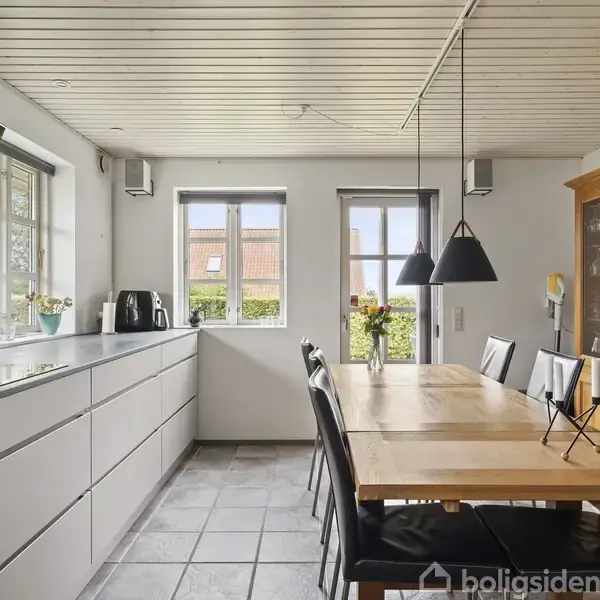Køkken med lang spisebord i lyst træ står centralt. Seks sorte stole omringer bordet. To vinduer og skydedør slipper naturligt lys ind. Enkel modern belysning hænger over bordet.