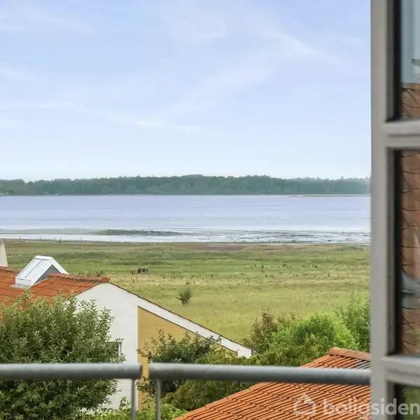 En udsigt fra en balkon viser et fredeligt landskab med røde hustage i forgrunden, en grøn eng i midten og en rolig sø i baggrunden under en klar himmel.