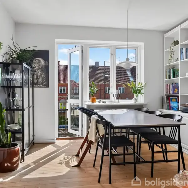 En spisestue har et mørkt bord og fire sorte stole. Store vinduer åbne mod en byudsigt. Reoler fyldt med bøger og planter omkranser rummet. En kaktus står ved siden af en glasreol.