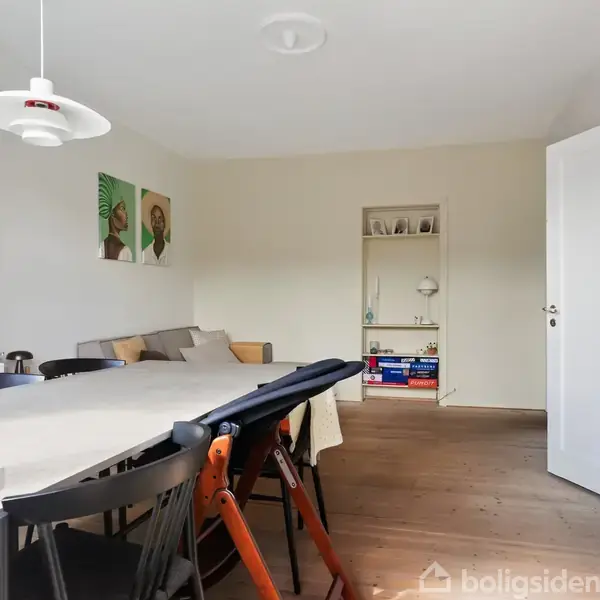 Et spisebord med stole står i et lyst rum ved siden af en hvid reol fyldt med bøger og planter. En åben dør fører til et tilstødende værelse med spil.