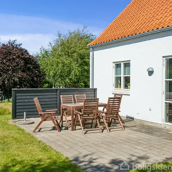 Terrassemøbler står på en flisebelagt terrasse foran et hvidt hus med rødt tegltag. I baggrunden er der grønne buske og træer under en klar blå himmel.