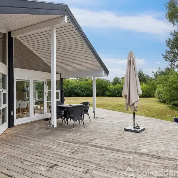 Træterrasse med et udendørs bordarrangement under en parasol ved siden af en moderne bygning med store vinduer. En stor græsplæne og buske omkranser området.