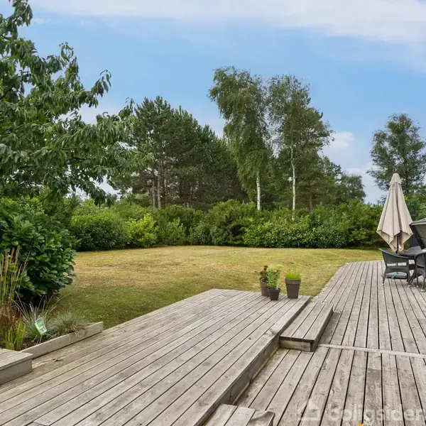 Træterrasse med tomme potteplanter står stille i en have. Græsplæne og buske omgiver området, mens nogle træer ses i baggrunden under en blå himmel.