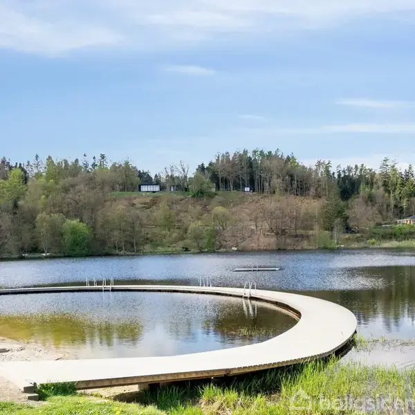 En rund træbro strækker sig over en rolig sø, omgivet af frodig skov og en blå himmel. Broen starter ved en lille sandstrand i forgrunden.