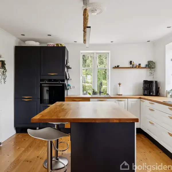 Et moderne køkken med en sort og træfarvet køkkenø i centrum. Rummet har hvide skabe, en ovn, dekorative planter, og hvide vægge. Store vinduer lader naturligt lys strømme ind.