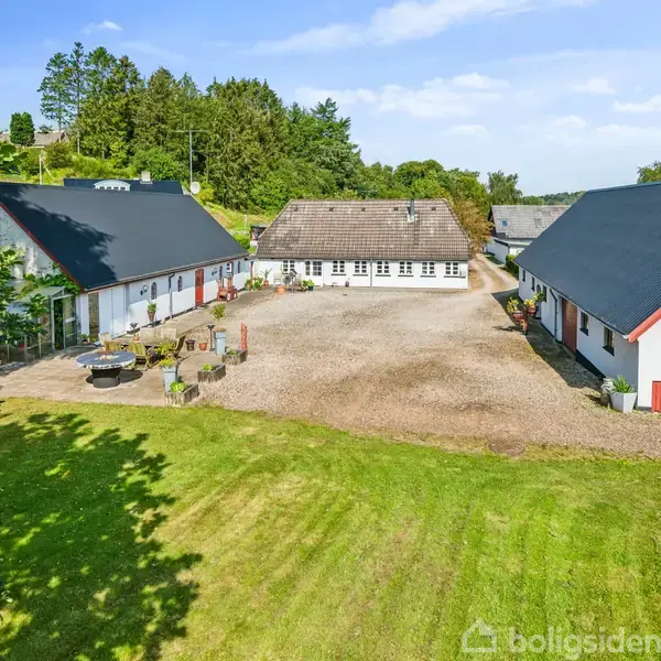 Et gårdkompleks består af tre hvidmalede bygninger med sorte tag, placeret omkring en grusbelagt gårdsplads. Foran bygningerne er der en grøn græsplæne, omkranset af træer.