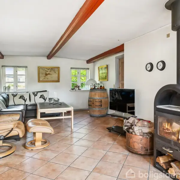 Pejs i hjørnet varmer stuen med lænestol og sofa på flisegulv. Fjernsynet står på et møbel ved siden af vinfad og vinduer med kraftige loftsbjælker.