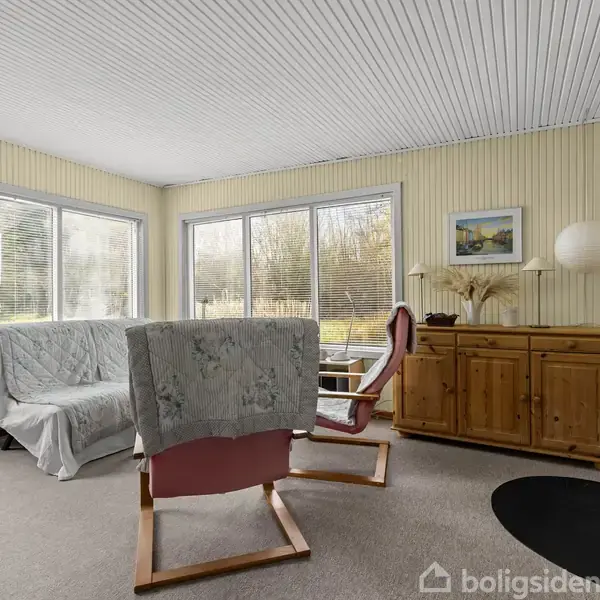 Stue med sofa og lænestol, dækket af tæpper, står foran store vinduer. En kommode med dekorative genstande og en brændeovn fuldender det hyggelige, lyse rum.
