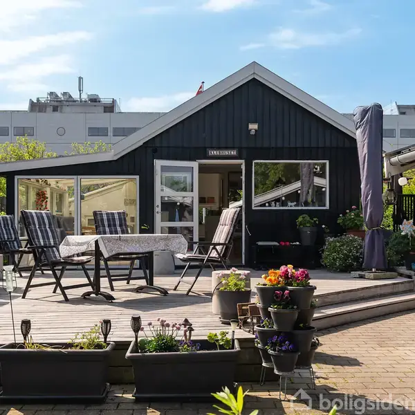 Sort, moderne sommerhus med stor terrasse, hvor der er placeret et havebord og stole. Omgivet af blomsterkrukker og grønne planter. Bygninger i baggrunden. Indgangsskiltet siger "TORVEGÅRD".