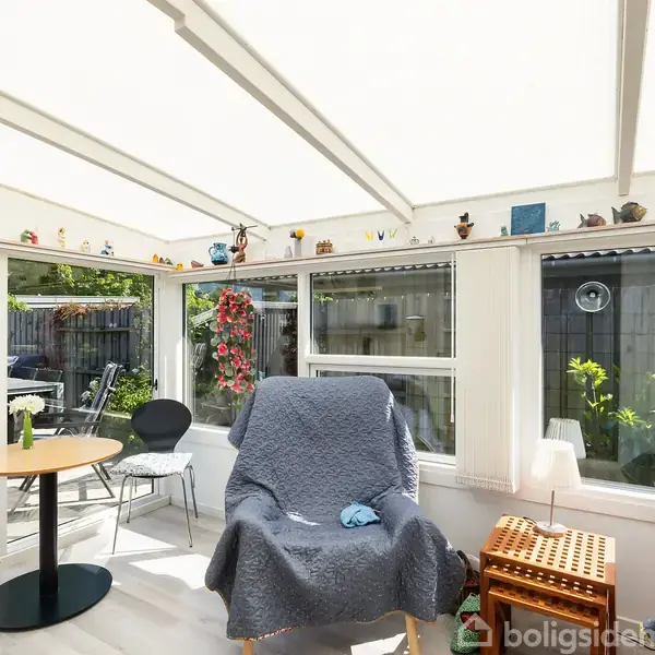 To polstrede stole dækket af grå tæpper står i en solrig udestue. Omringet af store vinduer viser udsigten en terrasse med havemøbler og grønne planter udenfor.