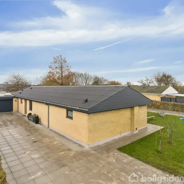 Hus i gule mursten med sort tag står stille. Indkørsel og legeplads i forhaven; træer og hække omgiver det. Skyfri blå himmel over boligområde.