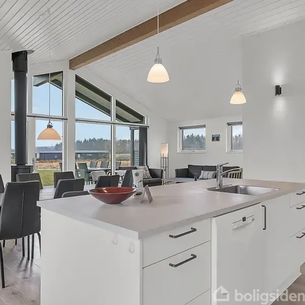 Køkkenø med vask, placeret i et lyst, moderne åbent rum. Et spisebord med stole står tæt på store vinduer med udsigt til grønt område. Pendellamper hænger fra loftet.