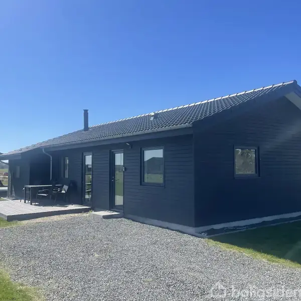 Mørkt træhus står stille. Terrasse med havemøbler på gruset grund omgivet af grønt græs. Solrig himmel giver lysklar kontekst.