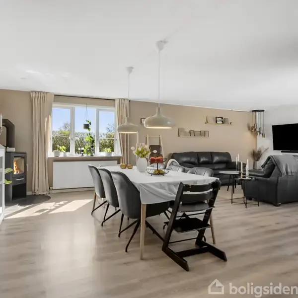 Stue med spisebord dækket med dug og blomster, omringet af stole. Sofaer står foran en tændt brændeovn og TV. Store vinduer lader lys strømme ind.