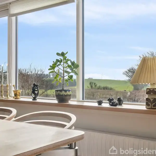 Et bord med tændte lys står ved siden af et stort vindue. Udenfor ses en grøn eng og træer. Vinduet har pynt som planter og en lampe på karmen.