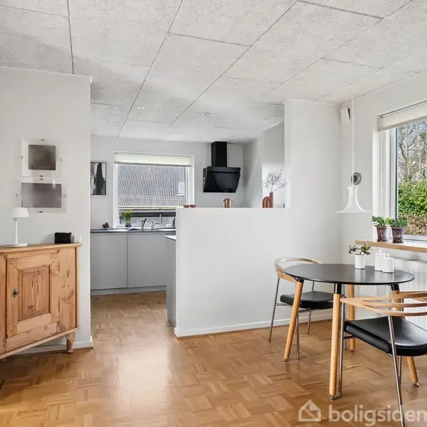 Et rundt spisebord med fire stole står i en lys spisestue. Bordet er placeret nær et vindue med udsigt til en have. Til venstre ses en åben dør til et køkken.