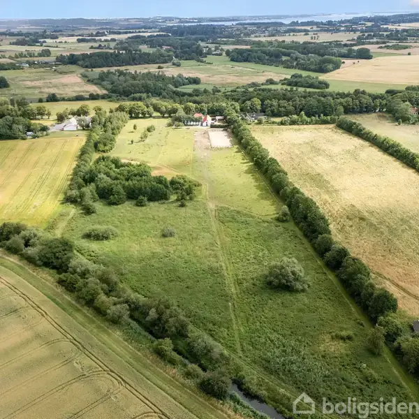Landskab set fra luften viser et stort, grønt landbrugsområde med marker og spredte træer. I baggrunden er der små bygninger omkranset af skov og bakker.