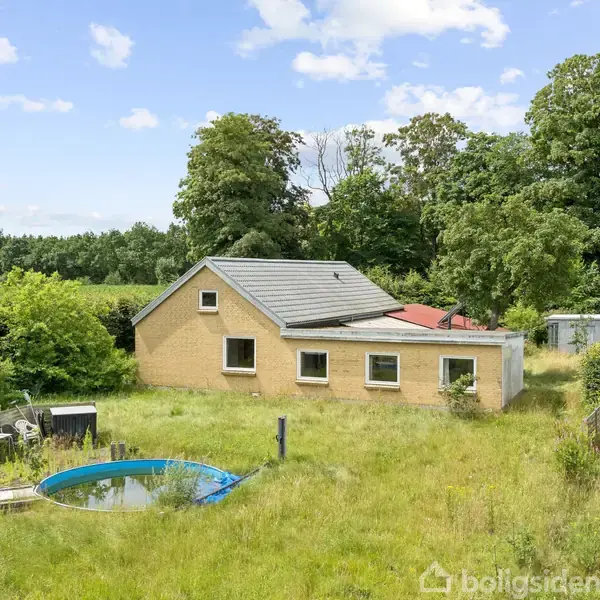 Et gult murstenshus står stille i en frodig have med højt græs. En lille, rund, blå pool er i forgrunden omgivet af træer og buskadser.