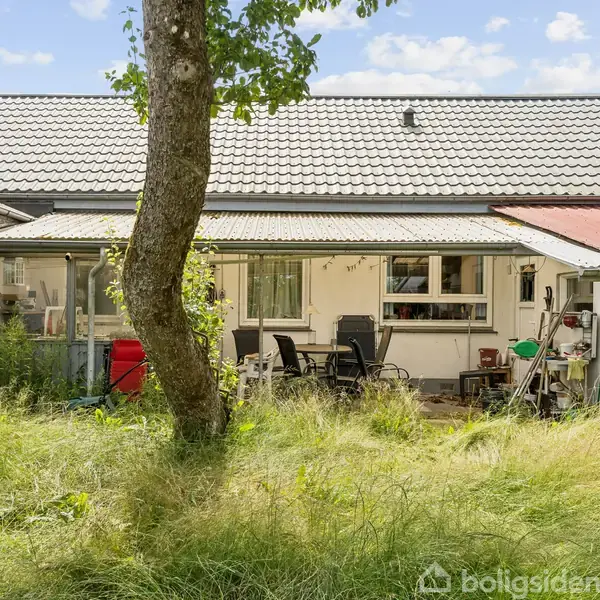 Hus med hvide vægge og et gråt tag, omgivet af højt græs og træer, viser en terrasse med havemøbler og diverse redskaber.