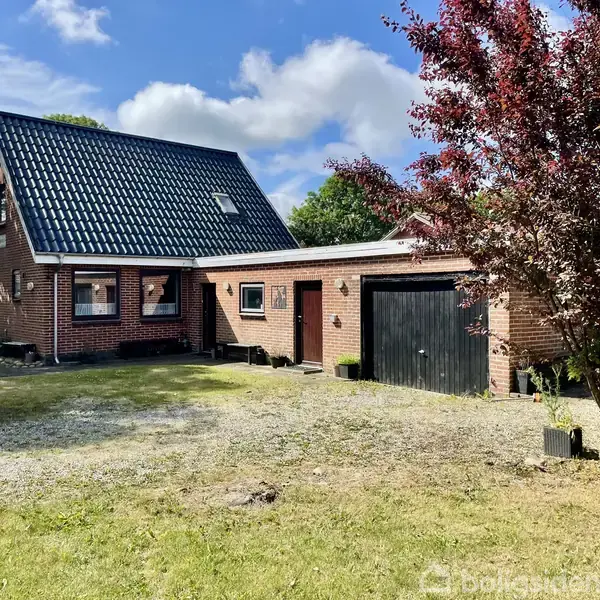 Et rødt murstenshus med sorte tagsten og tilhørende garage står stille i en have med græsplæne og buske. Himlen er blå med hvide skyer.