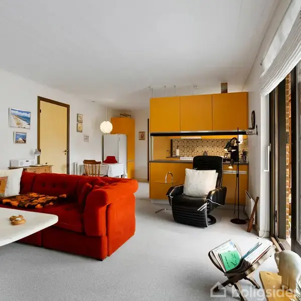 Rød sofa står i en stue med åbent køkken. Gule køkkenskabe ses i baggrunden. Store vinduer åbner ud til en have. Rummets indretning er retro med lamper og dekorative billeder.