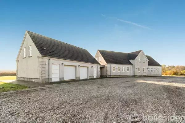 En stor hvid bygning med sort tag står stille på en grusbelagt gårdsplads. Omgivet af åbne marker under en klar blå himmel.