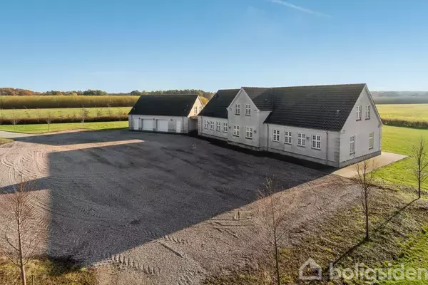 En stor, hvid bygning med sort tegltag står på en grusbelagt gårdsplads. Den er omgivet af åbne marker og træer på en solrig dag.
