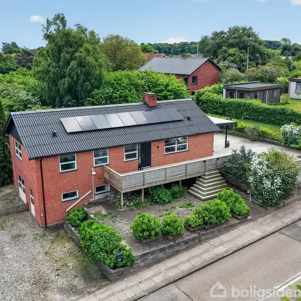 Hus med røde mursten og sort tag, placeret på en grøn grund. Solpaneler på taget. Træterrasse foran, muret trappe ved indgangen. Omgivet af træer og buske i roligt kvarter.