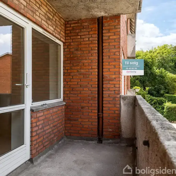 Balkon med murstensvæg, dør og til salg-skilt. Ved siden af en boligbygning, omgivet af grønne buske og træer med trapper, der fører ned til en sti.