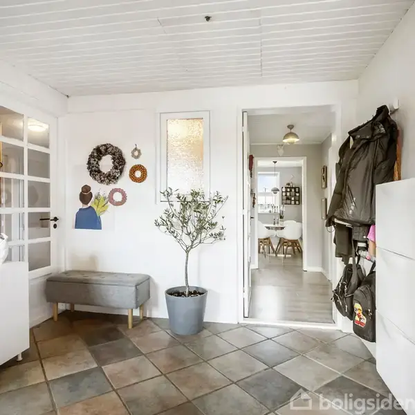 En entré med grå fliser på gulvet, en grå bænk, vægdekorationer, og et planter i potte. Til højre hænger jakker, og i baggrunden ses en spisestue.