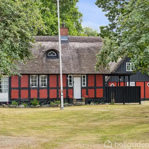 Bindingsværkshus med stråtag står stille i en have omgivet af grønne træer. Huset har røde og sorte vægge med hvide vinduer. En flagstang står foran huset.