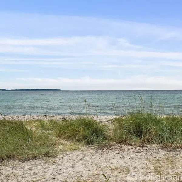Sandstrand foran roligt hav med bølgende græs i forgrunden. Himlen er klar med lette skyer i baggrunden. Ingen tekst til stede.